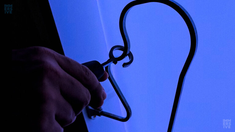 Close-up da mão de uma pessoa segurando o bastão com argola metálica enquanto tenta percorrer a barra do labirinto elétrico sob iluminação azul néon.