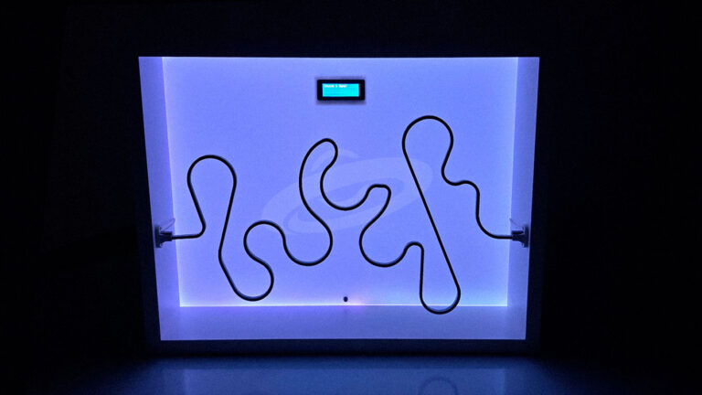 Labirinto elétrico aceso no escuro com backlight LED, exibindo o percurso sinuoso de metal e a tela digital no centro superior.