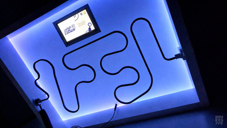 Labirinto elétrico visto de baixo, iluminado com luz LED azul ao fundo, destacando a haste de metal sinuosa e uma pequena tela digital na parte superior no centro.